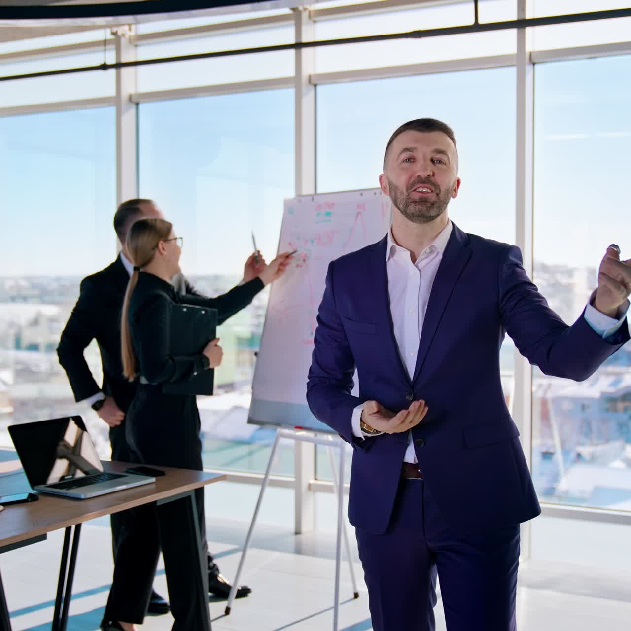 Portrait of a businessman talking on camera. Confident entrepreneur in blue costume speaking about his success in the light office with business people working