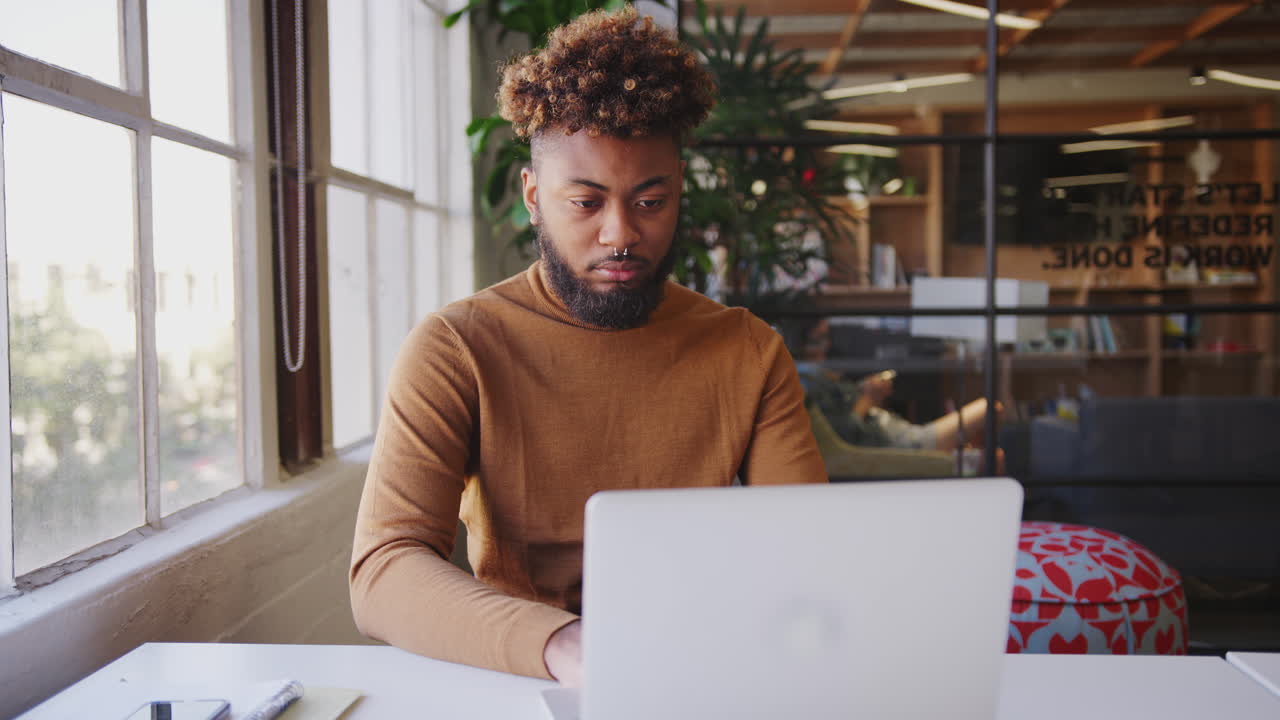 Millennial black male blogger using laptop  at a desk by the window in a creative office, close up