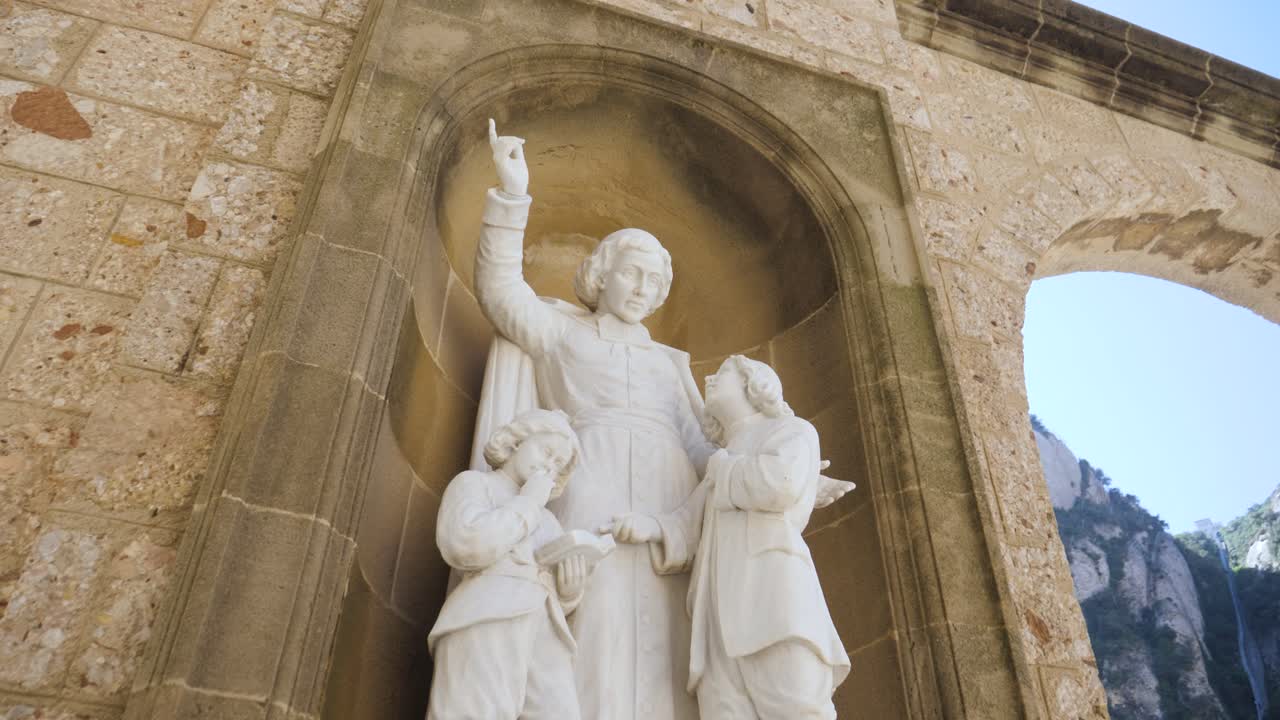Un primo piano di una scultura in marmo bianco raffigurante una figura santa che punta verso l'alto mentre insegna a due bambini piccoli. La statua è collocata all'interno di una nicchia di pietra tradizionale nel complesso dell'Abbazia di Montserrat
