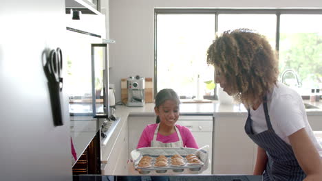 Happy-biracial-mother-and-daughter-baking-in-kitchen-taking-cakes-out-of-oven,-slow-motion