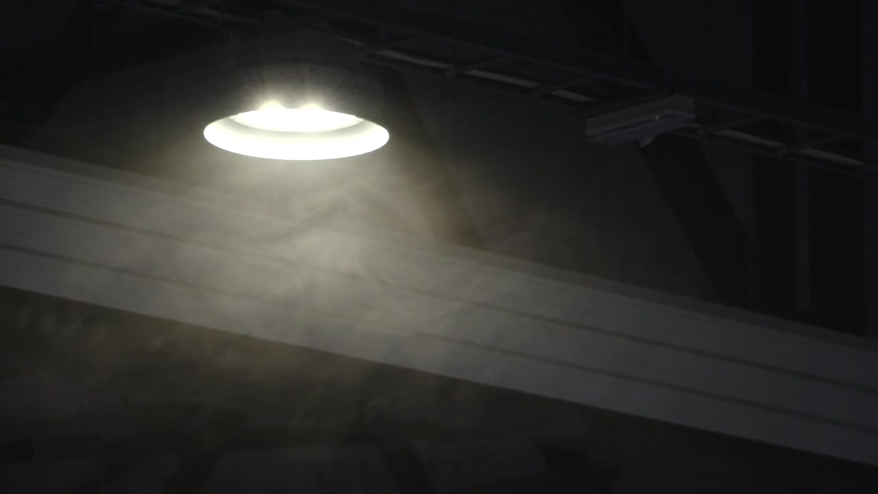 Una lámpara de techo brillante ilumina a través del polvo y la niebla en un interior oscuro, creando una atmósfera sombría con haces de luz dramáticos y sombras profundas.