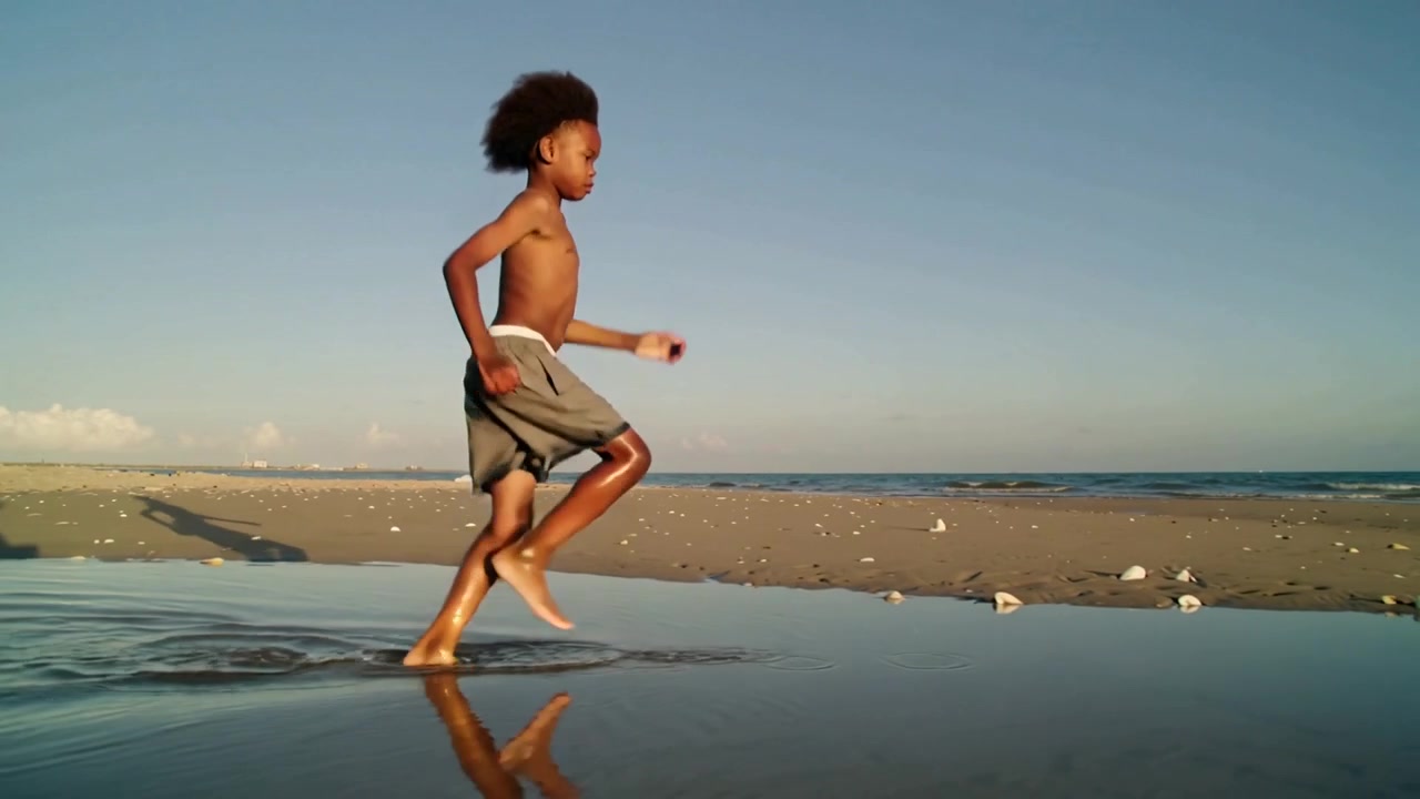Løbende dreng uden trøje i grå shorts træder ned i en tidevandspøl på stranden og laver plask, spejling.