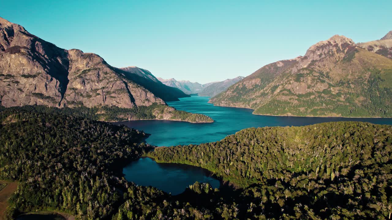 A breathtaking drone shot capturing the extreme scale of the Patagonian wilderness. Features towering, rugged mountain peaks, a deep glacial fjord, and untouched forest surrounding a small