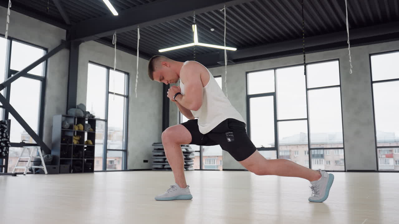 Athlete stretching legs in gym studio performing side lunge with clasped hands, wearing white tank top and black shorts, large windows and overhead lights highlighting warmup focus and strength