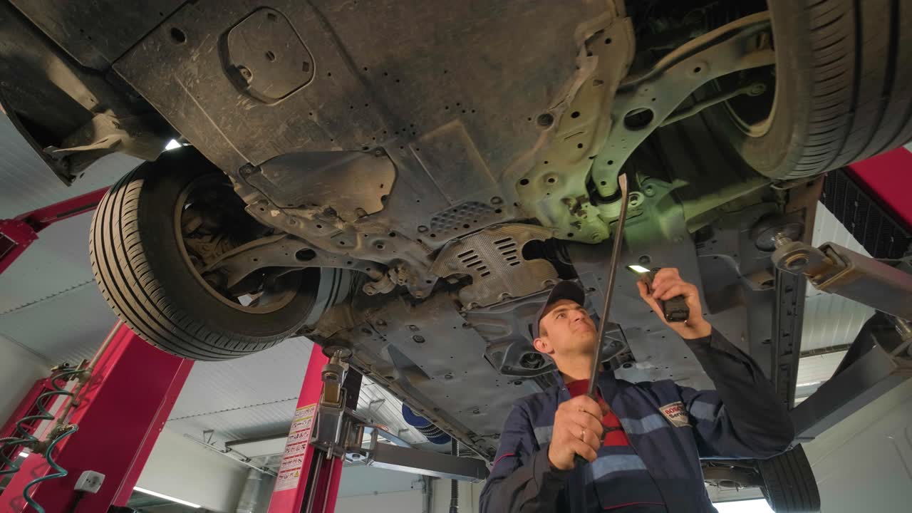 Young car mechanic at repair service station inspecting car wheel and suspension detail of lifted automobile. Bottom view.