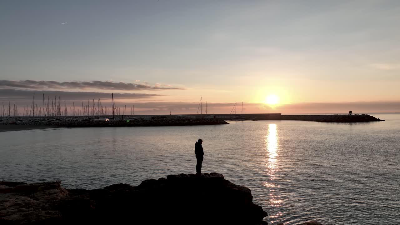 Amazing drone view of unrecognizable traveler standing on edge of rocky cliff near white houses and admiring picturesque sea view at sunset