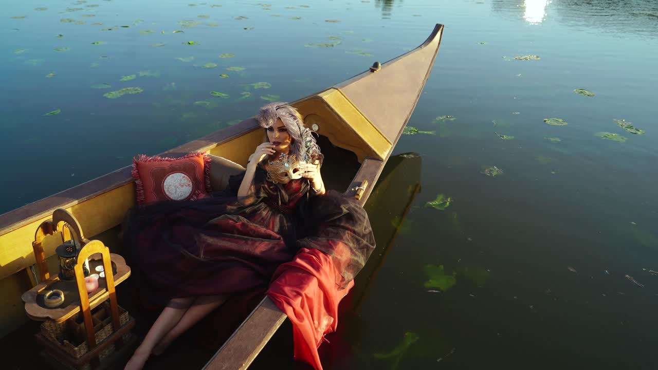Woman dressed in traditional costume with carnival mask lying in a boat in Venice. Attractive young sensual romantic woman floating in gondola in the evening.