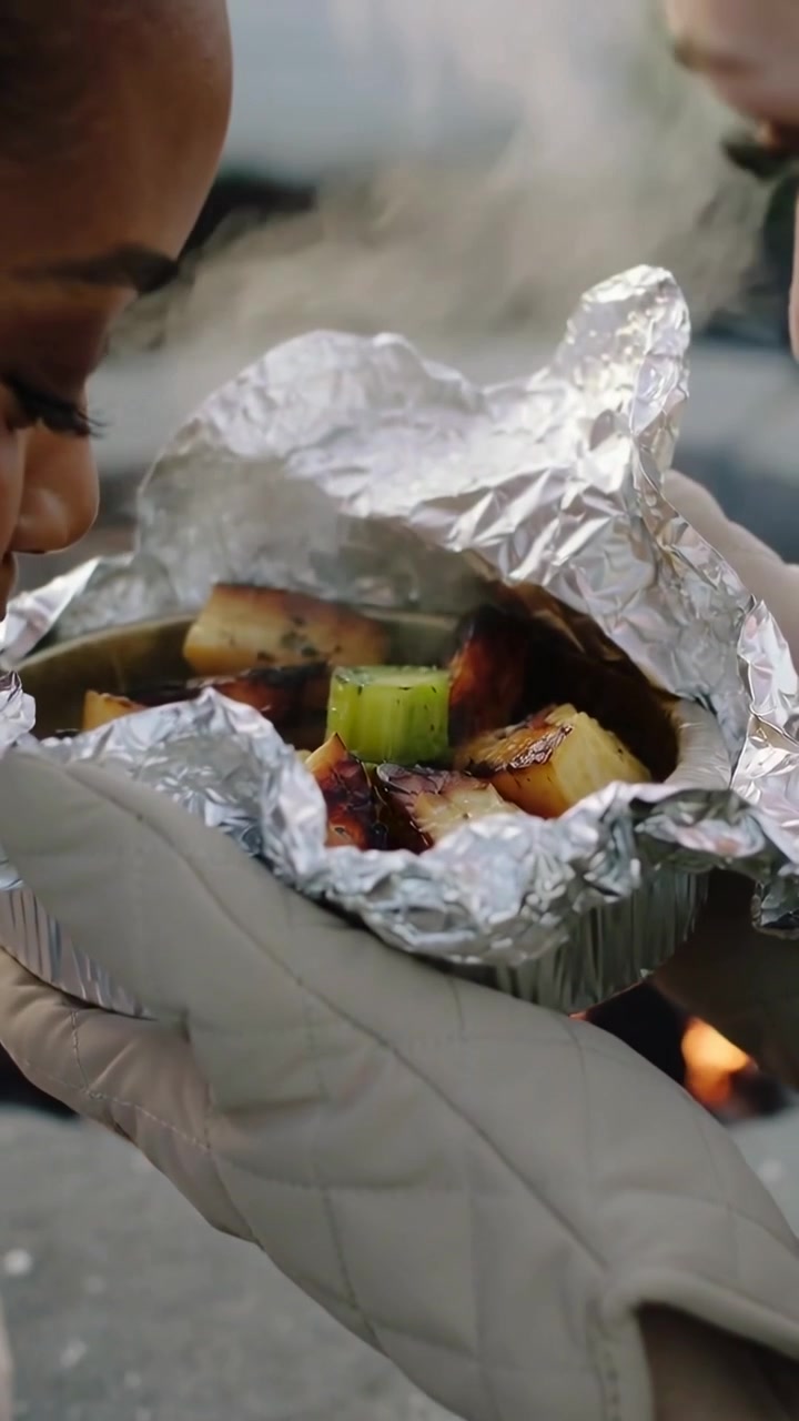 Video verticale: Un cuoco, stimolato dal calore, con guanti da forno trapuntati apre un vassoio di alluminio sopra una fossa di mattoni, entrambi annusano.