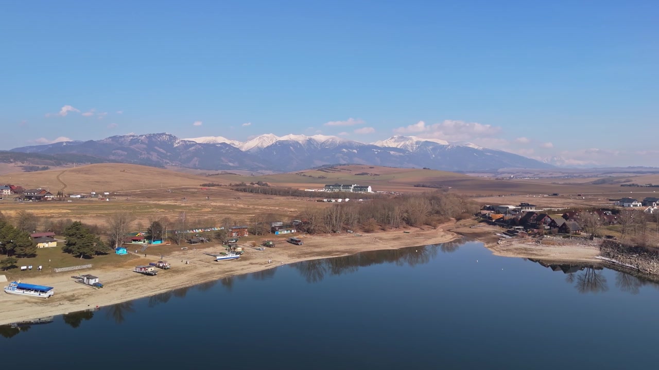 Il bacino idrico di Liptovský Trnovec e Liptovská Mara in Slovacchia con barche, cottage sulla riva e le montagne Tatra innevate riflesse sull'acqua calma in un panorama invernale ripreso da drone.
