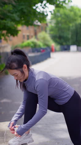 Vertical-Video-Of-Young-Woman-Tying-Laces-On-Training-Shoe-Before-Exercising-Running-Along-Urban-Street