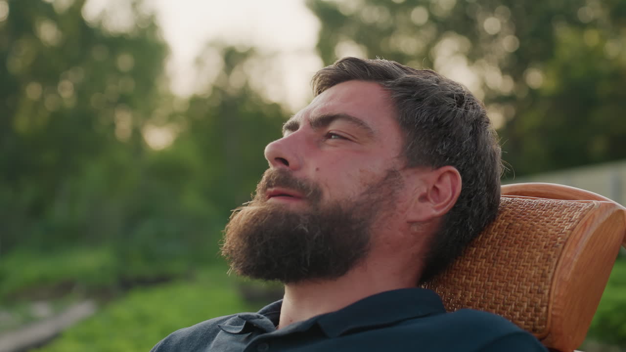close up side view man sipping hot coffee from clear glass mug while relaxing in wicker rocker chair on lawn near white fence at sunset with soft bokeh foliage and steam rising peaceful moment