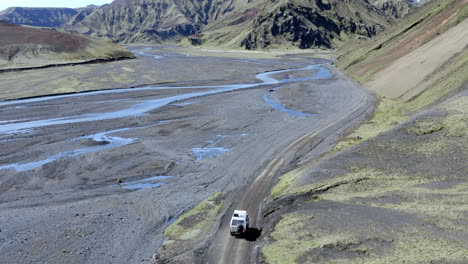 Coche-4x4-Conduciendo-Por-Un-Camino-De-Grava-Gris-Entre-El-Río-Glaciar-Y-Las-Laderas-De-Las-Montañas-Cubiertas-De-Musgo-Verde-En-Islandia