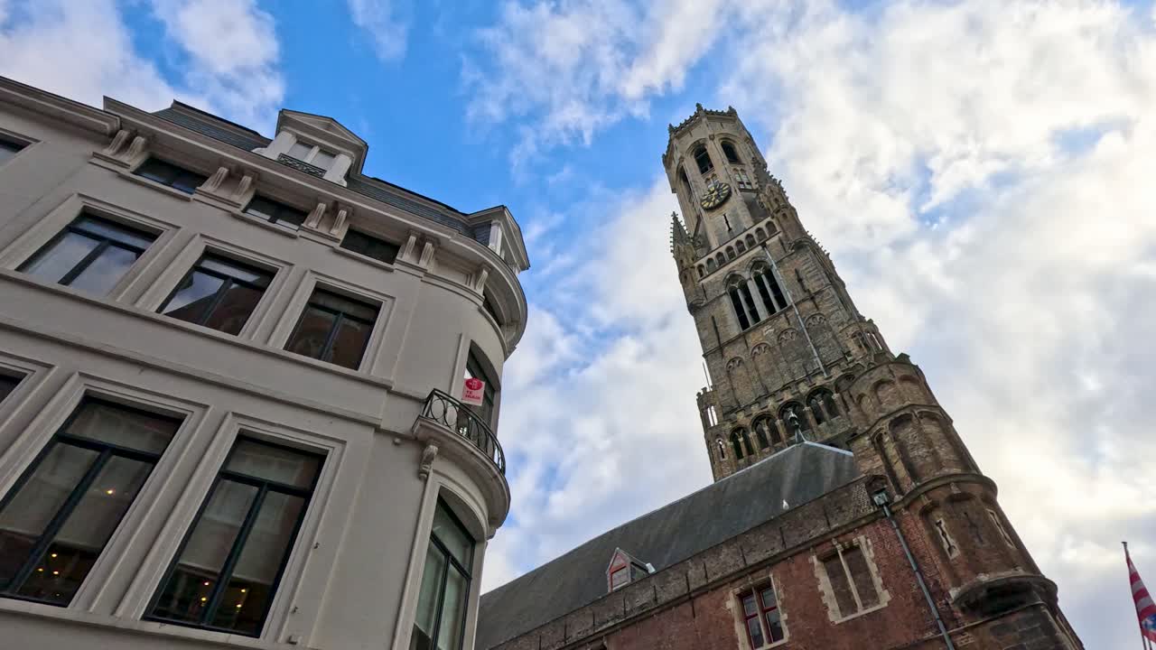 Camera tilts up from street, revealing Bruges medieval Belfry tower and historic buildings in daylight