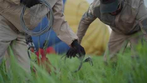 female crew member kneeling on lush grass coiling steel cable during hot air balloon preflight preparation near colorful partially inflated envelope under evening sky beside teammate in gloves