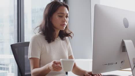 Millennial-Asian-woman-sitting-at-an-office-desk-using-a-computer-and-drinking-coffee,-waist-up