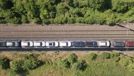 top-down aerial view of a freight train transporting vehicles along a railway track. the green locomotive leads a series of wagons carrying neatly arranged cars through a green landscape.