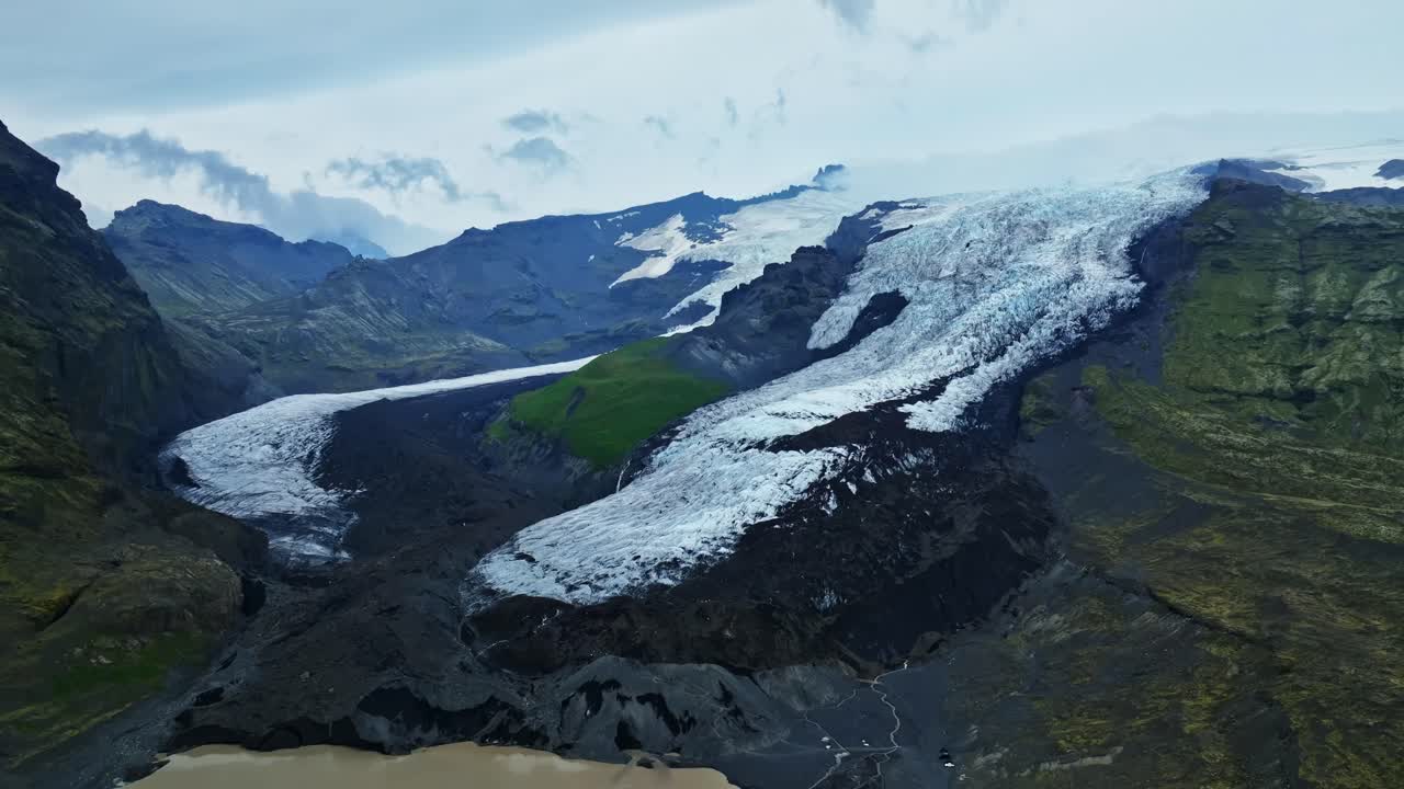Højtliggende dronepanorama over en Vatnajökull-udløbsgletsjer, der flyder gennem robuste vulkanbjerge mod en mælkehvid smeltevandslagune i Island