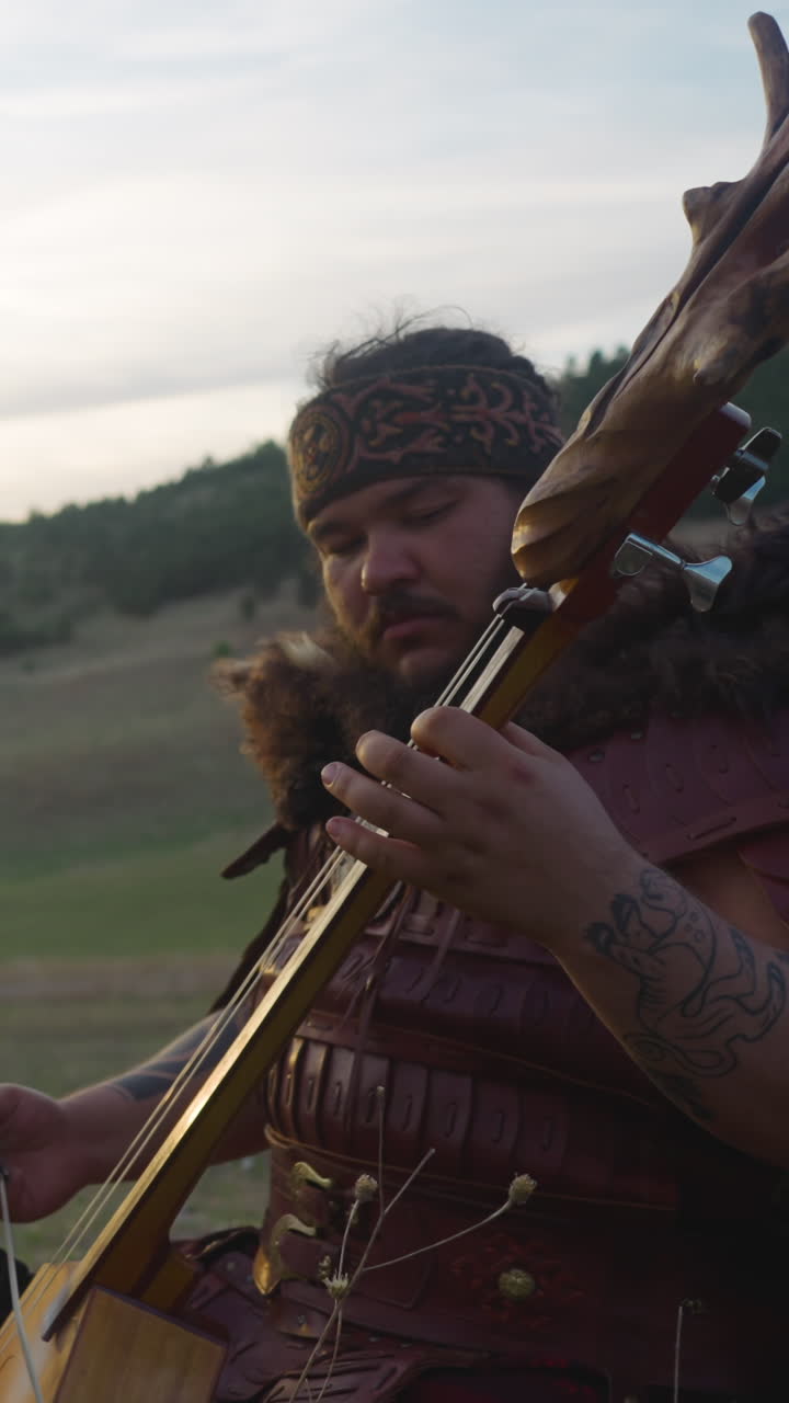 Large man in shaman costume plays ikili sitting in mountain valley at sunrise light. Siberian folks music at festival. Protecting old traditions