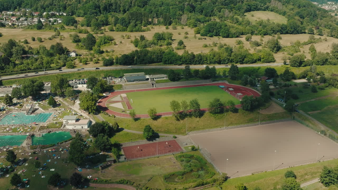 Establishing drone shot captures a large multi-sport complex in the Odenwald forest region of Germany. Features a green soccer field, red running track, vibrant outdoor swimming pools