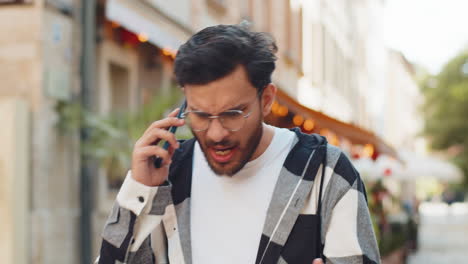 angry hispanic young man tourist arguing while talking on smartphone while walking on city street