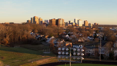 Slow-descent-from-a-downtown-city-skyline-to-a-football-field,-stadium-light-in-foreground