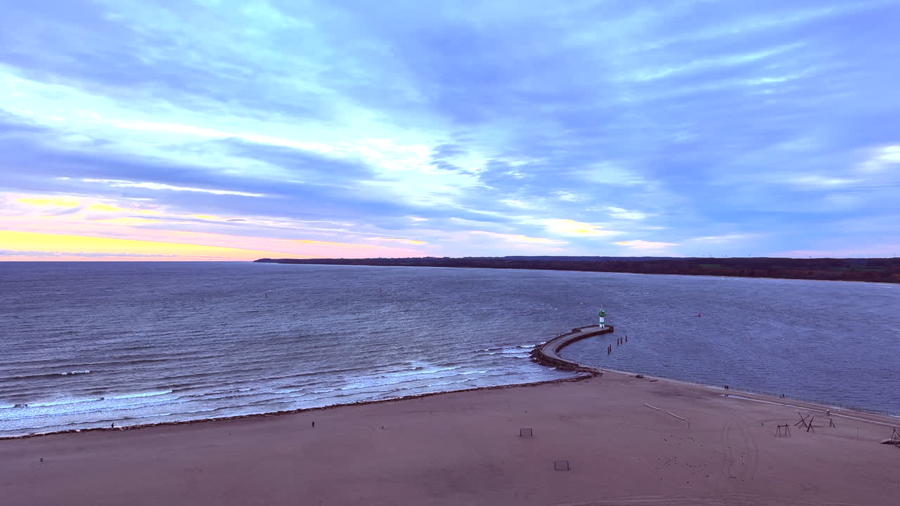 The video appears to be filmed in Travemünde and shows a windy coastal scene at the harbor entrance. A curved pier leads out to the green-and-white lighthouse at the breakwater