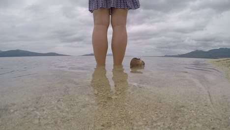 un video de 60 fps de un coco a la deriva lentamente al lado de una chica de pie en el borde de las aguas