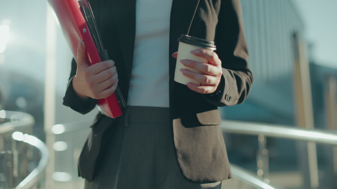 Close up partial view of banker walking while holding file folder with coffee cup in hand under bright sunlight with iron railing reflecting modern outdoor work environment