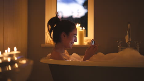 woman enjoying a relaxing bubble bath at night, using her phone.