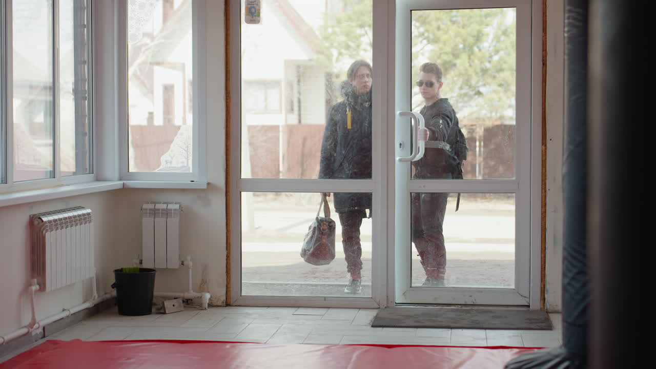 Two guys wearing dark jackets walks into a glass door of training hall, one adjusting sunglasses, one holding bag, visible through window, preparing to enter gym with red mats and punching bag inside