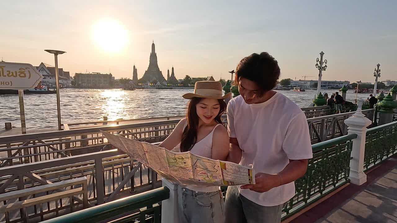 Coppia di turisti sorridenti studia la mappa della città su un molo sul fiume al tramonto dorato, foto a mano libera con inquadratura media