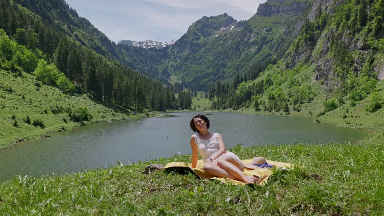 Brunette haired woman lying on picnic blanket during sunny day. Dolly forward shot. Green mountains with lake in valley. Swiss beauty in nature.