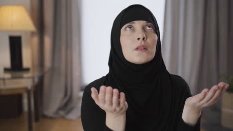 young muslim lady in hijab sitting with raised hands, looking up and shaking head. eastern woman in black hijab praying at home. religion, traditional culture.