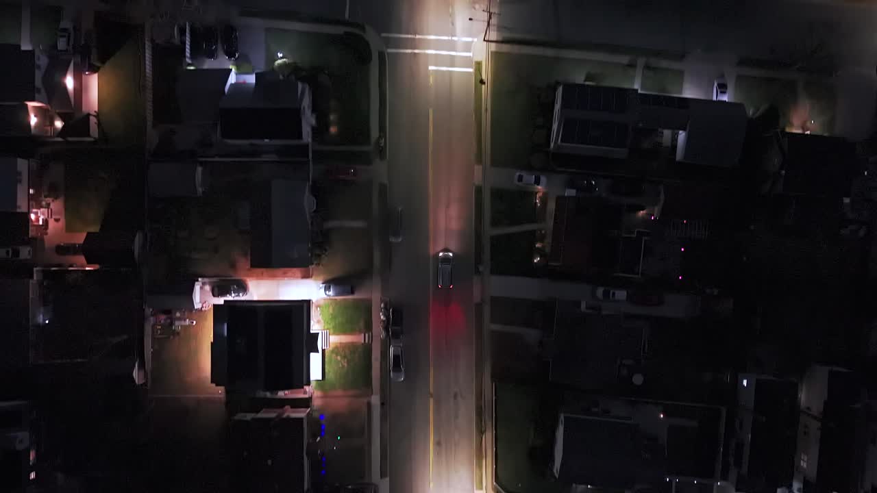 Vista aérea de drone diretamente para baixo, acompanhando um único carro por um bairro suburbano de Long Island, Nova York, à noite, capturando postes de luz brilhantes em uma tomada cinematográfica estilo vigilância.