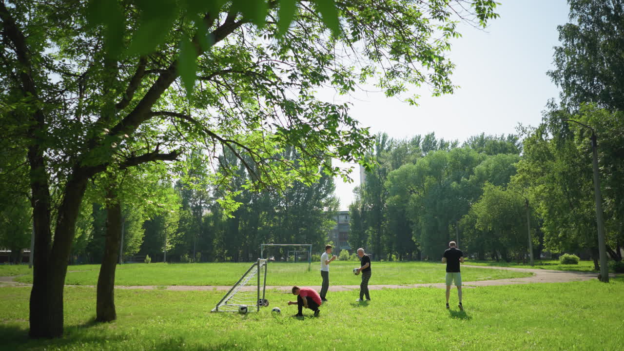 A family is doing outdoor exercise with son in red ties his shoelaces near a goalpost while his grandfather playfully hits his grandson's head with a ball