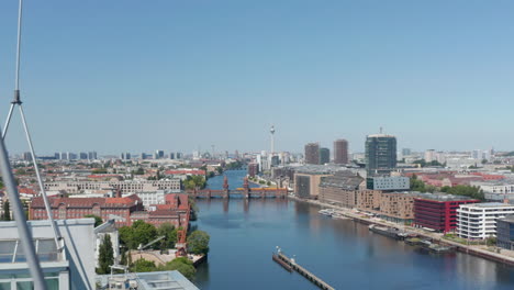 Fly-over-top-of-office-buildings,-reveal-of-historic-Oberbaum-bridge-with-train-crossing-Spree-river.-Fernsehturm-TV-tower-in-distance.--Berlin,-Germany