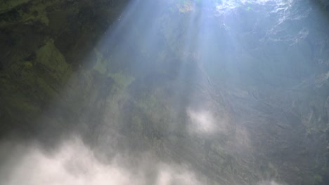 Pan-upward-with-sunbeam-shining-down-rocks-covered-with-green-plant-to-the-crack-on-the-ceiling-of-a-cave