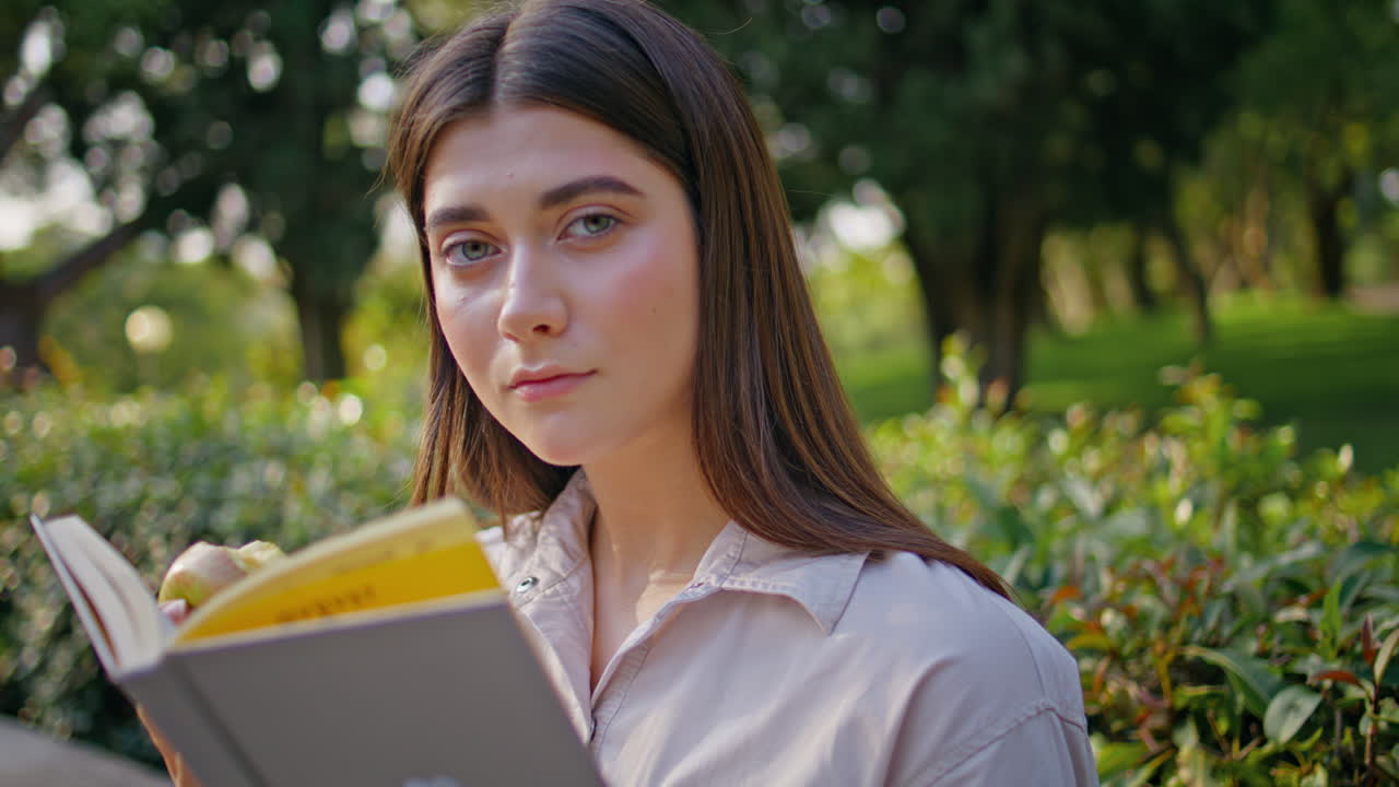 Ritratto di studentessa concentrata a leggere un libro in un giardino verde e soleggiato. La lettrice si sta godendo il momento.