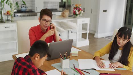 Asiatischer-Vater-Nutzt-Smartphone-Und-Laptop-Bei-Der-Arbeit-In-Der-Küche,-Während-Sohn-Und-Tochter-Schularbeiten-Erledigen