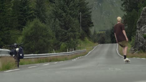 Young-caucasian-male-skateboarder-cruising-down-a-desolate-road,-away-from-camera,-in-the-middle-of-nowhere-in-Norway,-scandinavia