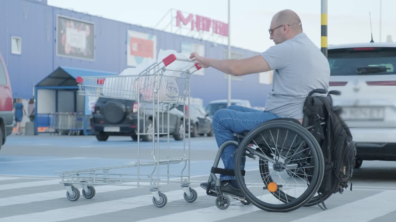 hombre con discapacidad en silla de ruedas empujando el carrito delante de sí mismo en el estacionamiento del supermercado