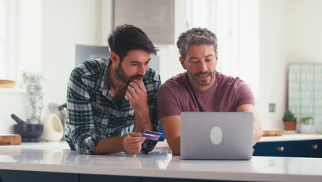 pareja de hombres del mismo sexo con tarjeta de crédito usando una computadora portátil en casa para reservar vacaciones o comprar