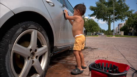 Feliz-Divertido-Niño-Latino-Mexicano-Sin-Camisa-Con-Pantalones-Cortos-Lavando-El-Auto-Familiar-Con-Un-Paño-Azul-En-Un-Día-Cálido-Y-Soleado