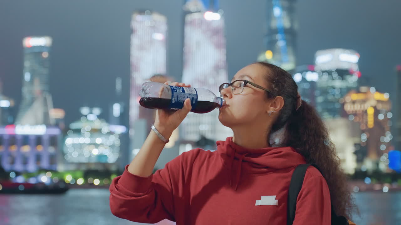Young woman drinking water by skyline, bottle in hand and red hoodie, sipping beside illuminated river with city lights reflecting relaxed nighttime pause, frames suggest jogger, commuter, traveler