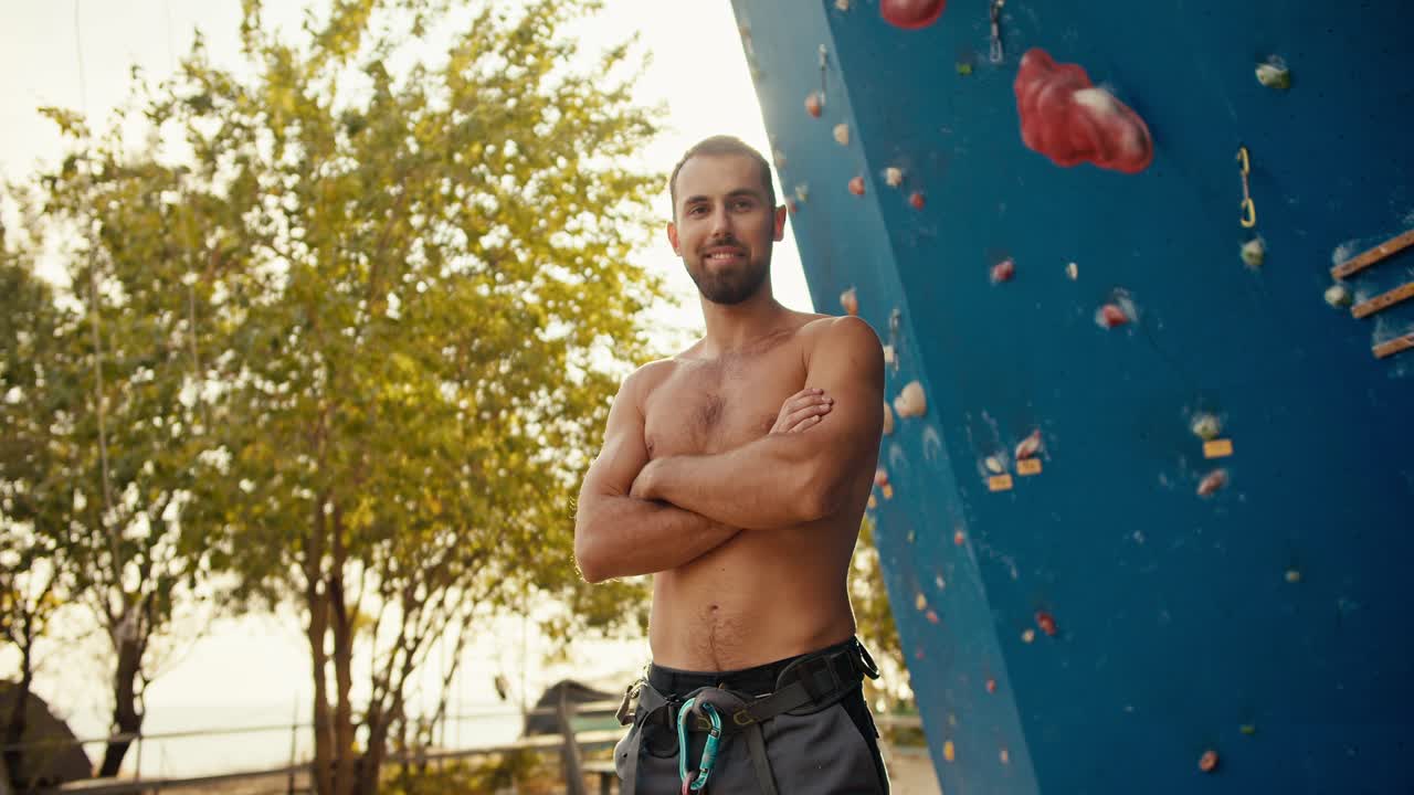 retrato de un feliz chico moreno con barba que dobla los brazos en su pecho y mira a la cámara cerca de una pared de escalada azul en un soleado día de verano. hombre experimentado scala con torso desnudo posando y mirando a la cámara junto a su pared de escaladura en verano