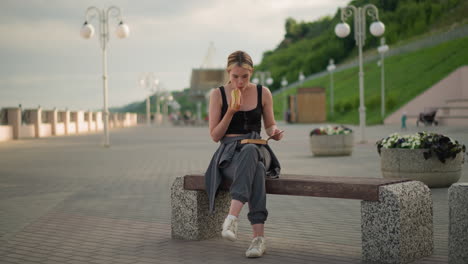 woman seated outdoors with crossed legs, holding a book in one hand while eating snacks with the other, she continues reading, with a blurred background featuring a lamp post, and people walking