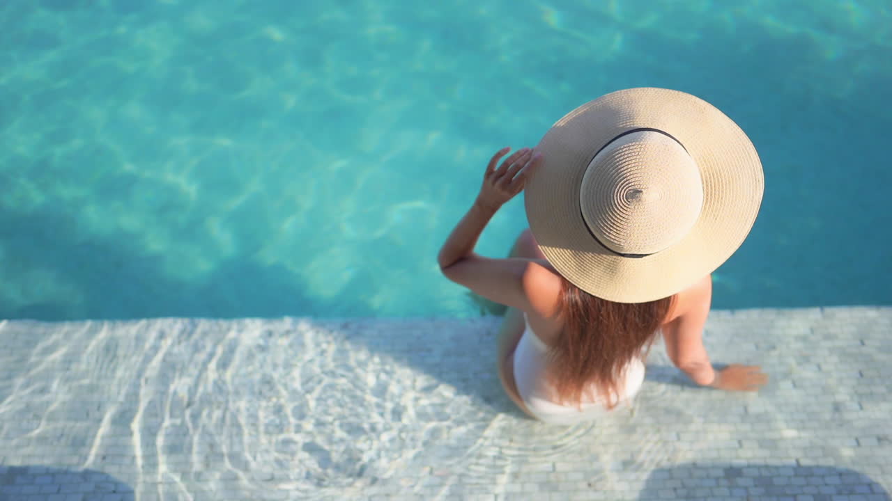 Back of Sexy Woman in Swimsuit and Floppy Summer Hat by Swimming Pool on Sunny Tropical Day