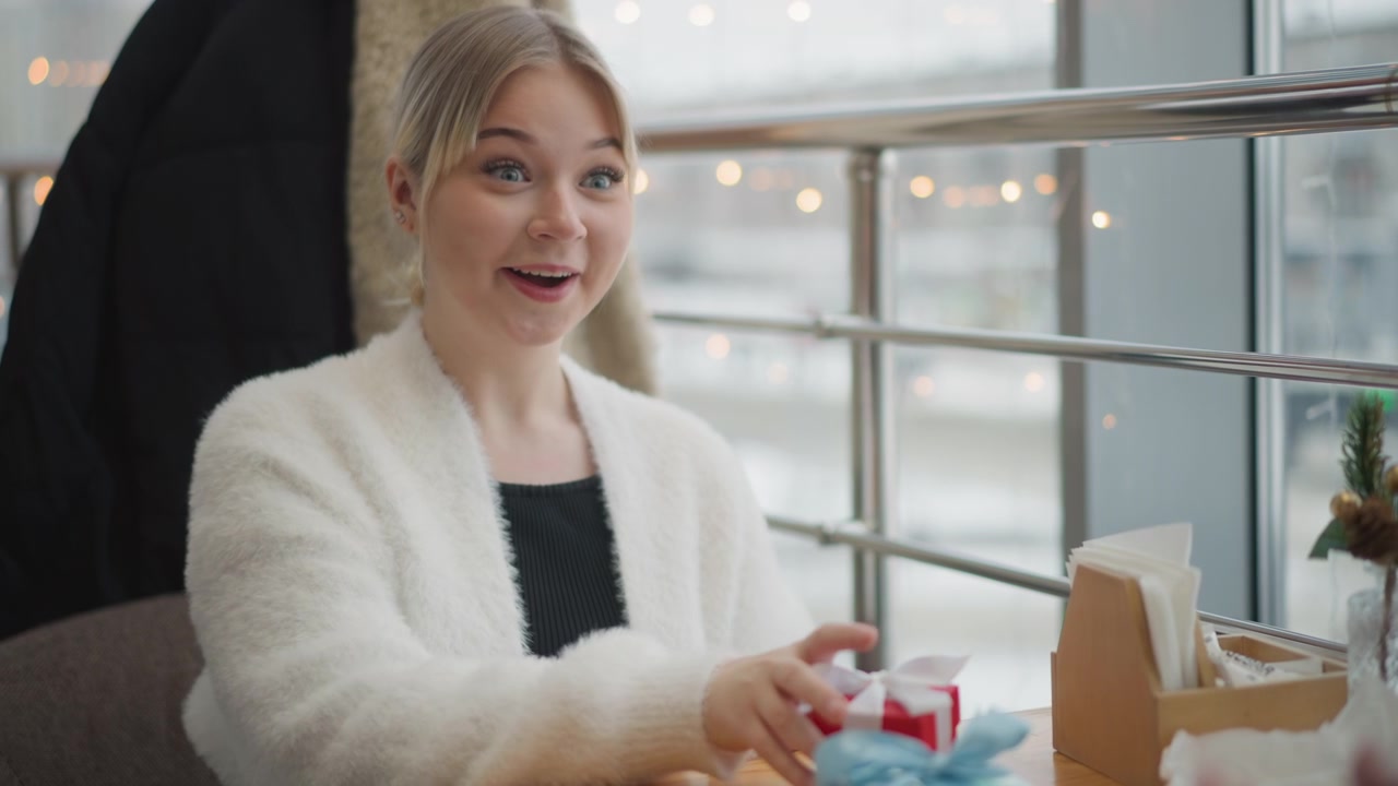 Donna bionda con giacca bianca pelosa seduta a un tavolo di caffè, chinata in avanti per presentare una piccola scatola regalo rossa verso un compagno con un viso sorridente, postura aperta e luce naturale soffusa vicino alla finestra.