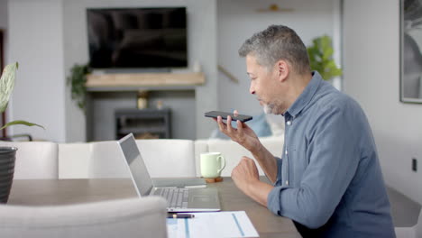 Älterer-Mann-Mit-Gemischter-Abstammung,-Der-Am-Tisch-Mit-Dem-Smartphone-Spricht-Und-Einen-Laptop-Benutzt,-Von-Zu-Hause-Aus-Arbeitet,-Zeitlupe