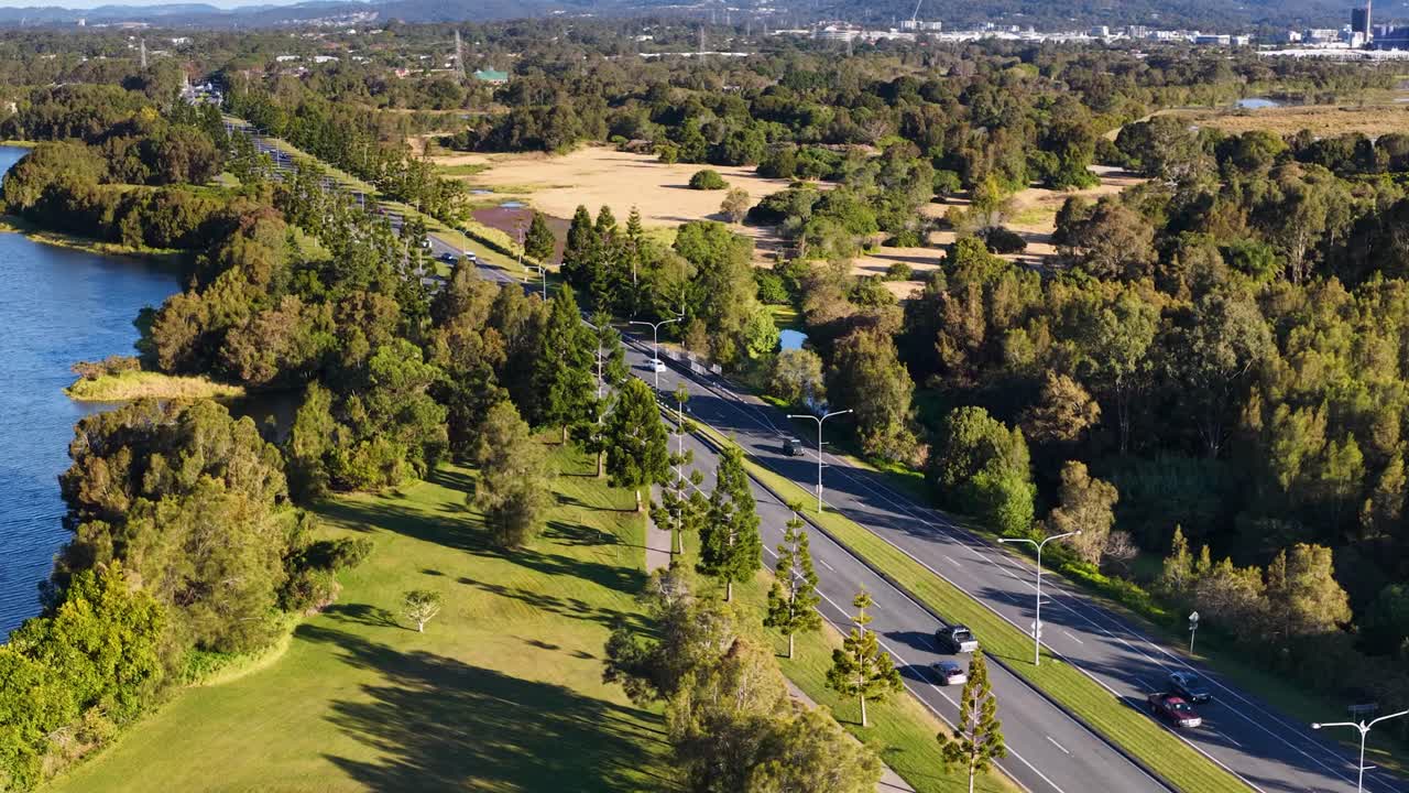 Drone footage captures light vehicle traffic moving along a winding road beside a river and lush greenery in Gold Coast, Australia, under bright daylight